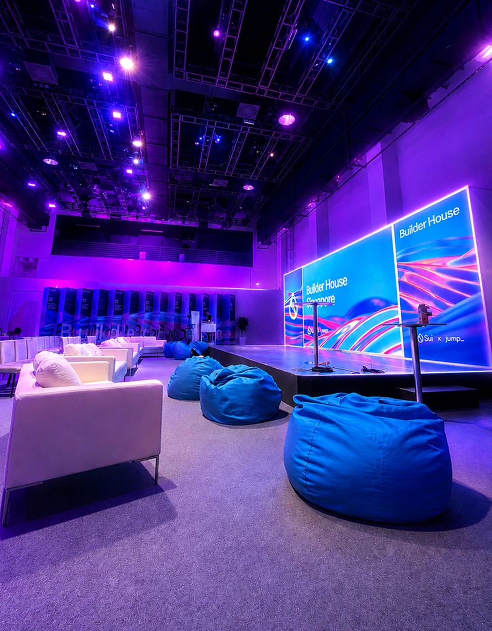Modern tech conference stage setup in Singapore featuring illuminated LED video wall, overhead truss lighting, and bean bag lounge seating
